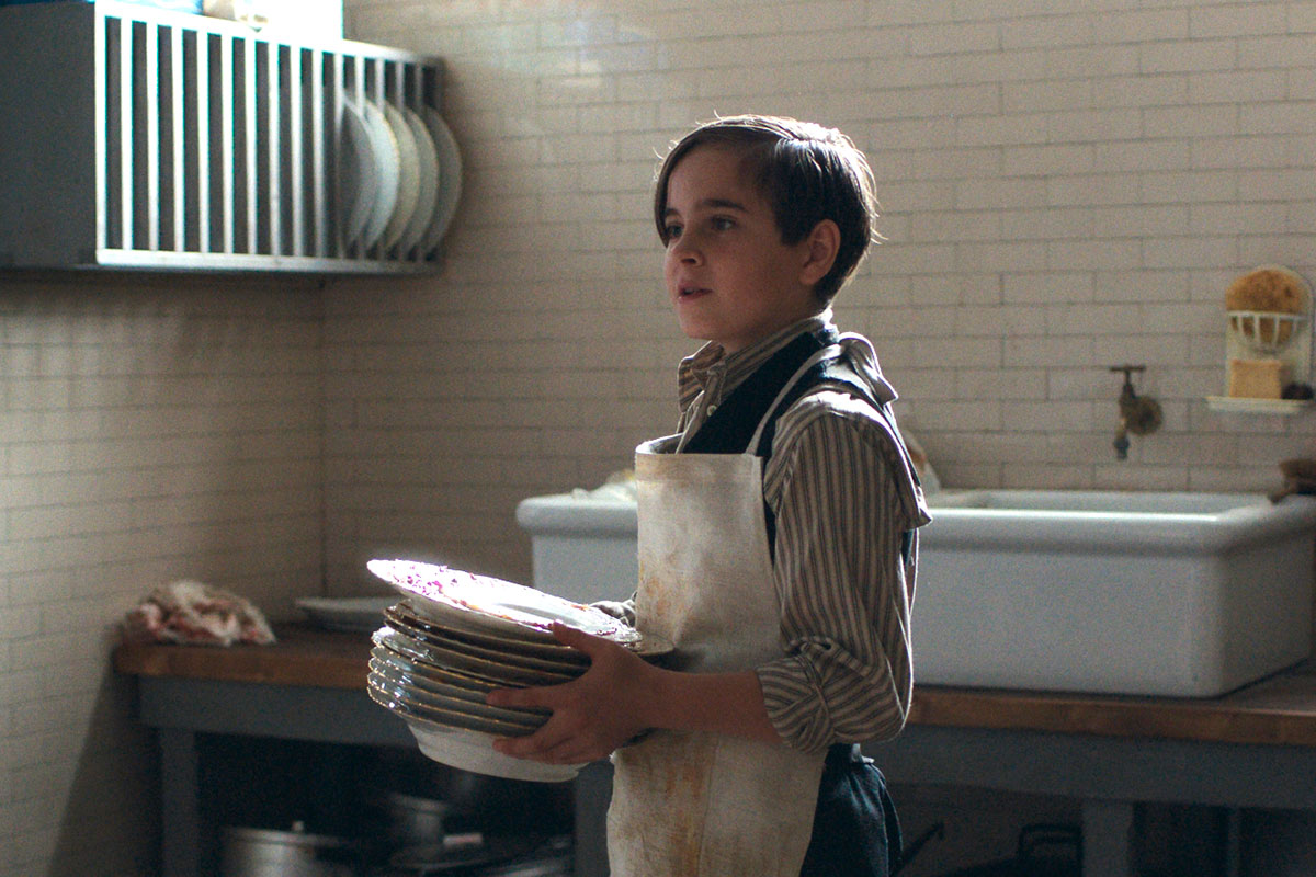 Georg steht mit Schürze in der spärlich beleuchteten Küche und trägt einen Stapel Teller