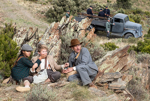 Die drei verstecken sich auf einem Hügel hinter einem Baumstumpf, im Hintergrund ein Militärfahrzeug mit Soldaten
